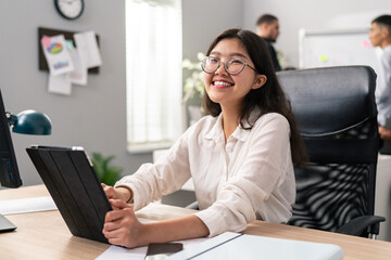 A smartly dressed woman with glasses and a white shirt works in the claims and customer service department of a large corporation, holding a tablet in her hands, replying to emails, smiling - Powered by Adobe