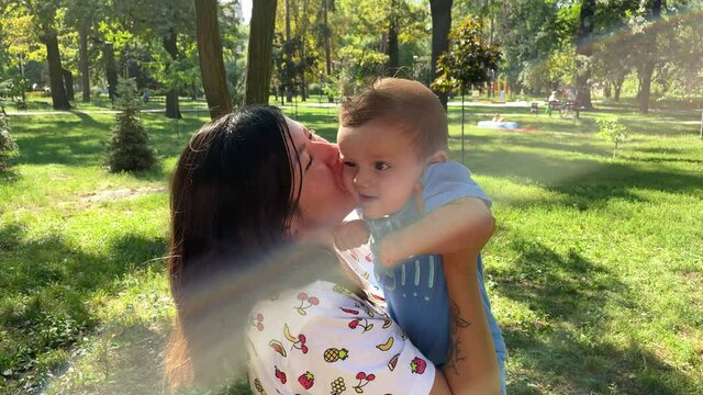 a young beautiful black-haired caucasian mother holds her smiling baby son in her arms and kisses him in a meadow in a green park in the rays of the sun