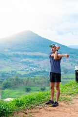 Portrait of an Asian male runner in a blue shirt and hat standing warm up in preparation for a jogging run. It is located on a high cliff with beautiful mountains in the background.