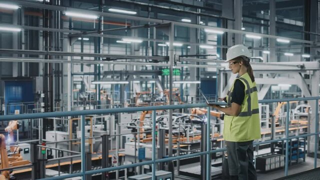 Female Car Factory Engineer in High Visibility Vest Using Laptop Computer. Automotive Industrial Manufacturing Facility Working on Vehicle Production with Robotic Arms. Automated Assembly Plant.