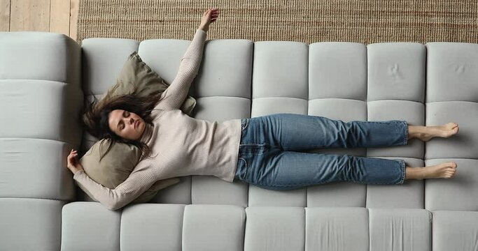 Above top full length view exhausted young woman sleeping on cozy couch, enjoying break pause time at home. Millennial lady feeling tired after hard working day, fallen asleep on cushions at home.