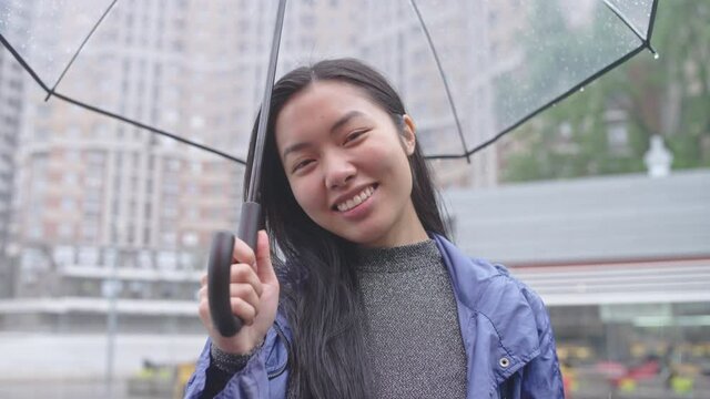 Happy asian woman smiling, enjoying rain, walking city with transparent umbrella