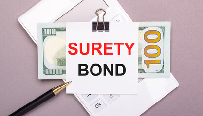 On a gray background, a white calculator, a pen, banknotes and a sheet of paper under a black paper clip with the text SURETY BOND. Business concept