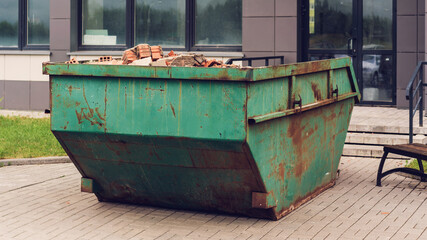 Fototapeta na wymiar Green rusty metal container with orange brick debris against building