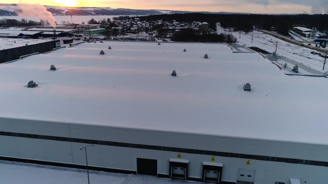 Huge building of production plant workshop with snow on flat roof and rising up steam in winter evening aerial view