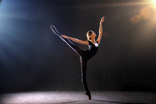 ballerina in tight-fitting suit is dancing on black background on pointe shoes, silhouette is illuminated by sources of color.