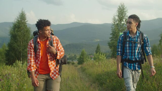 Positive handsome multiracial male travelers with backpacks checking position by using portable magnetic compass, looking for right direction while trekking in mountains at sunset.