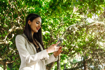 White young businesswoman using smartphone near the trees. She is smiling. Horizontal copy space