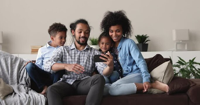 Happy young african american father holding smartphone in hands, recording video or posing for selfie photo with joyful wife and little adorable kids siblings, resting together on comfortable sofa.