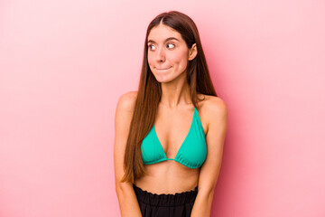 Young caucasian woman wearing bikini isolated on pink background confused, feels doubtful and unsure.