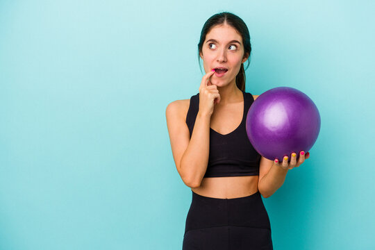 Young caucasian fitness woman holding a ball isolated on blue background relaxed thinking about something looking at a copy space.
