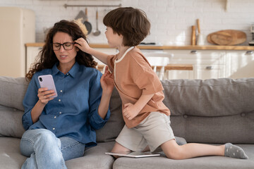 Hyperactive bored little kid distract young mother businesswoman work on mobile phone from home....