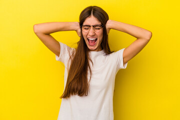 Young caucasian woman isolated on yellow background covering ears with hands trying not to hear too loud sound.