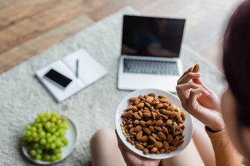 cropped view of woman eating almonds near blurred gadgets and fresh grape on floor.