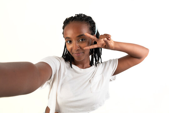 Self photo of a woman smiling with two finger in her face, looking at camera. Selfie picture. White background. African-Ethiopian black woman.