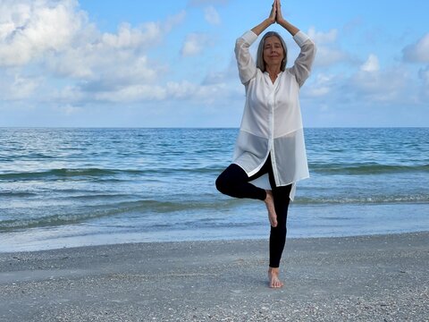 Senior Woman Relaxing In A Standing  Yoga  Pose On The Beach With The Sea And Sky As Background