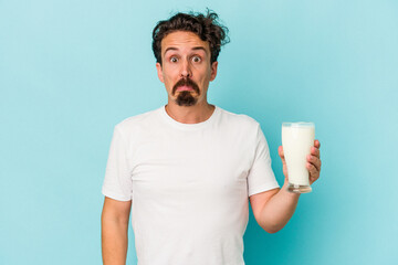 Young caucasian man holding a glass of milk isolated on blue background shrugs shoulders and open...