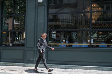 Successful stylish man in formal black suit walking on the street near dark-green wall. High quality photo