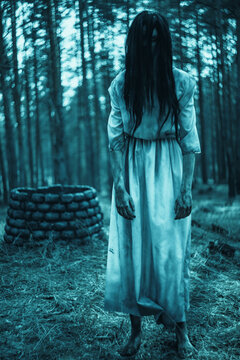 Girl in image of scary zombie stands in dark forest near stone well.