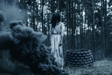 Girl in image of scary zombie walks in dark forest among smoke.