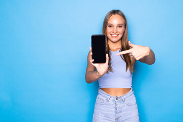 Fototapeta premium Portrait of a beautiful young woman in sweater showing blank screen mobile phone isolated over blue background