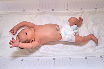 A newborn baby in diapers on a white changing table with a ruler with a Moro reflex - obrazy, fototapety, plakaty
