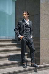 Handsome man in black formal suit and yellow sunglasses standing on the stairs and keeping his hands in his pockets. High quality photo