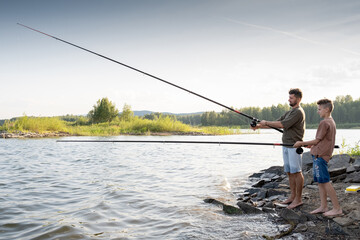 Teenage boy and his father fishing together by lake on summer weekend