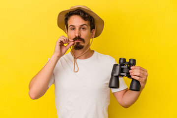 Young caucasian man holding binoculars isolated on yellow background with fingers on lips keeping a secret.