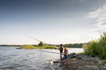 Active man and teenage boy with rods fishing by lake on summer weekend