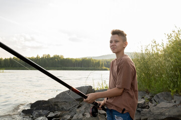 Contemporary teenage boy with long rod fishing alone by lake on summer weekend