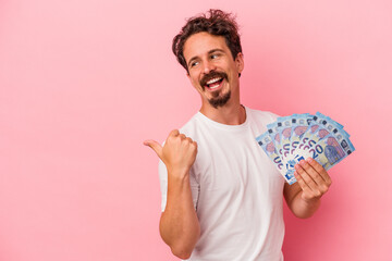 Young caucasian man holding banknotes isolated on pink background points with thumb finger away, laughing and carefree.