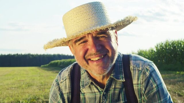 Smiling elderly farmer portrait. Agronomist wearing straw hat looking in camera with smile. Happy senior man with grey beard posing on plantation background. Sunny day, countryside concept.