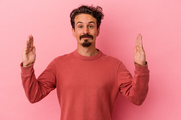 Young caucasian man isolated on pink background holding something little with forefingers, smiling and confident.