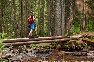 Fototapeta premium Traveler hiker with backpack crosses mountain river in Carpathian forest walking on bridge. Trip to summer Ukraine