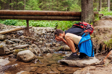 Traveler hiker with backpack washes face in mountain river in Carpathian forest by bridge. Tourist having rest