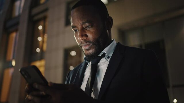 Online chatting. Stylish african american man in suit typing on cellphone, standing outdoors in evening, tracking shot