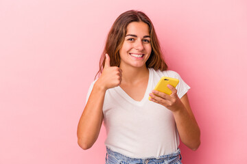 Young caucasian woman using mobile phone isolated on pink background  smiling and raising thumb up