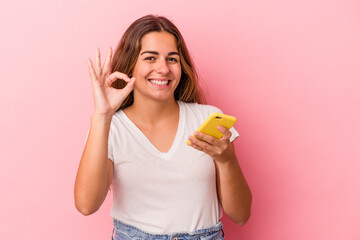 Young caucasian woman using mobile phone isolated on pink background  cheerful and confident showing ok gesture.