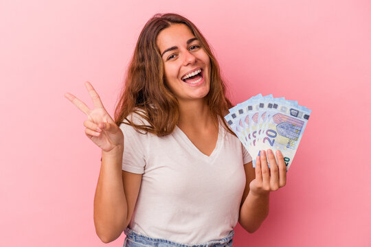 Young caucasian woman holding bills isolated on pink background  joyful and carefree showing a peace symbol with fingers.
