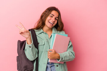 Young caucasian student woman holding books isolated on pink background  joyful and carefree showing a peace symbol with fingers.