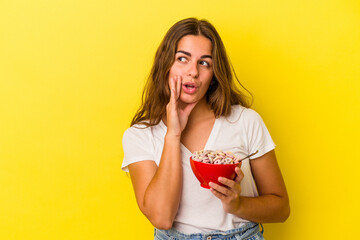 Young caucasian woman holding cereals isolated on yellow background  is saying a secret hot braking news and looking aside