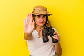 Young caucasian woman holding binoculars isolated on yellow background  standing with outstretched hand showing stop sign, preventing you.
