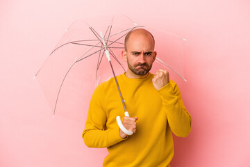 Young caucasian bald man holding umbrella isolated on pink background  showing fist to camera, aggressive facial expression.