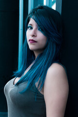 young woman with blue hair standing by the bus stop. looking towards the camera. serious latin girl posing standing by a wall in the street