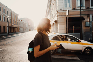 Fototapeta premium Young woman traveler with a backpack with a smart background in her hands is waiting for a taxi. Against the background of a city road with buildings.