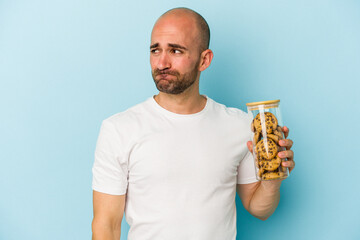 Young bald man holding cookies isolated on blue background  confused, feels doubtful and unsure.