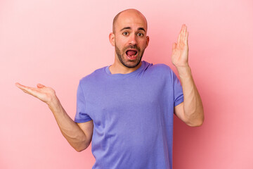 Young caucasian bald man isolated on pink background  holds copy space on a palm, keep hand over cheek. Amazed and delighted.