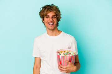 Young caucasian man with makeup holding popcorn isolated on blue background  happy, smiling and cheerful.