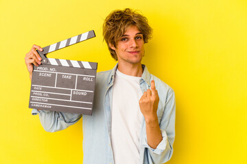 Young caucasian man with makeup holding clapperboard isolated on yellow background  pointing with finger at you as if inviting come closer.